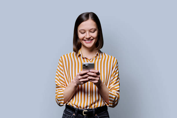 istock Young smiling woman using smartphone looking at phone in grey background 2187886264