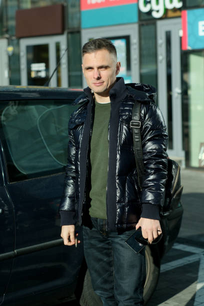A man casually dressed is poised by a parked car in a bustling city atmosphere.