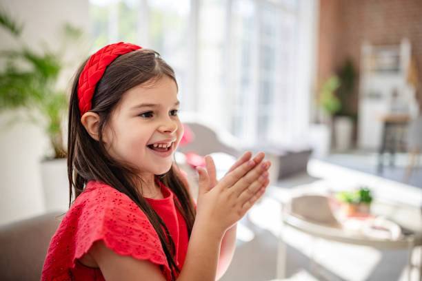 A young girl dressed in red sits on a couch, joyfully clapping her hands together, expressing excitement and happiness in a bright, modern home environment.