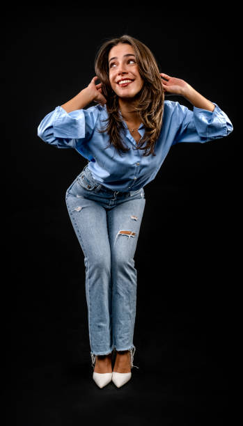 istock In a studio setting with a black background, a young brown-haired woman wearing a blue shirt and jeans is posing in an artistic style 2187920598