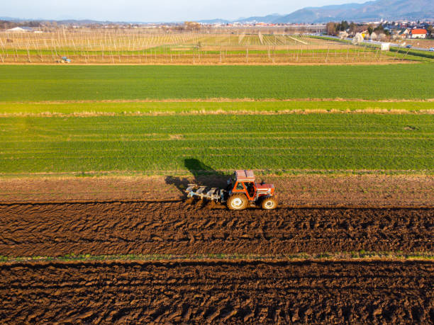 Aerial view of a tractor plowing a field with green grass and brown soil. The landscape features rows of crops and distant hills under a clear blue sky.