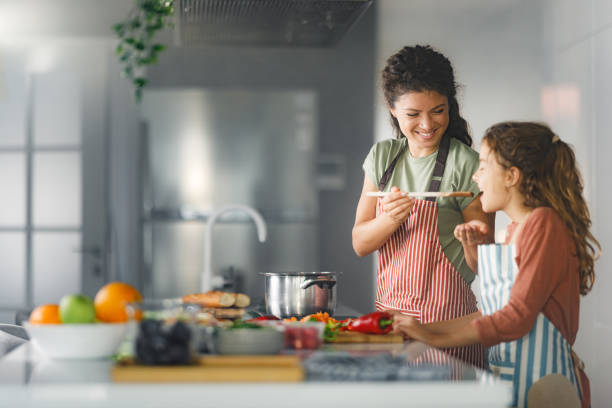 Try this sweetie! Happy single mother giving her daughter to taste the food while preparing a meal together in the kitchen. family dinners and cooking stock pictures, royalty-free photos & images