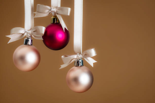 A festive arrangement of three hanging Christmas ornaments in metallic pink, rose gold, and burgundy hues, adorned with satin ribbons, set against a warm golden background.