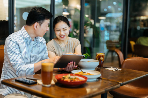 istock Two Asian mid woman and man using tablet in a cafe. Enjoying a relaxing moment in the using digital tablet application for online shopping. Emotional friends communication, laughing, looking at digital screen, sitting together. 2187977907