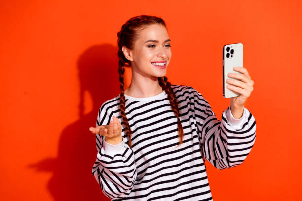 istock Young woman with red hair in striped pullover smiling and using smartphone against orange background showcasing modern casual style 2187982595