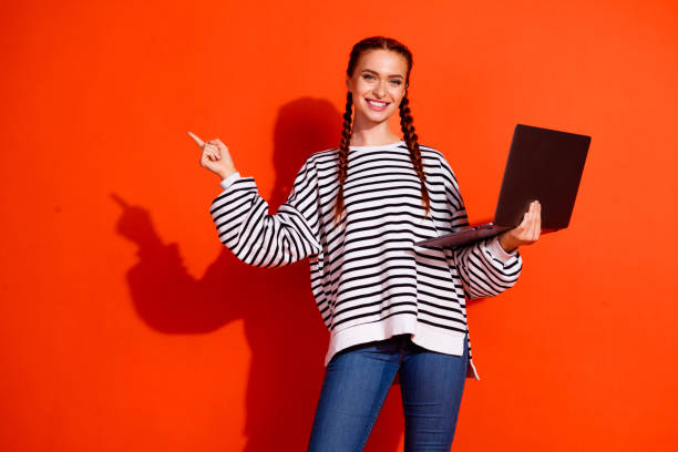 istock Cheerful woman in pullover holding laptop and pointing on vibrant orange background, highlighting casual style and advertising concepts 2187982630
