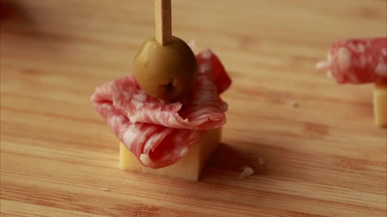 Preparing Party Appetizer with Salami, Old Cheddar, and Green Olives on Wooden Board Close-Up