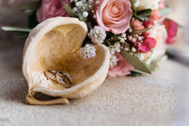 Close-up of two golden wedding bands delicately placed inside a natural walnut shell half, accompanied by a romantic floral arrangement featuring pink roses and delicate baby's breath flowers. The rustic, organic shell container creates an artful contrast with the elegant rings, while resting on a textured light surface.