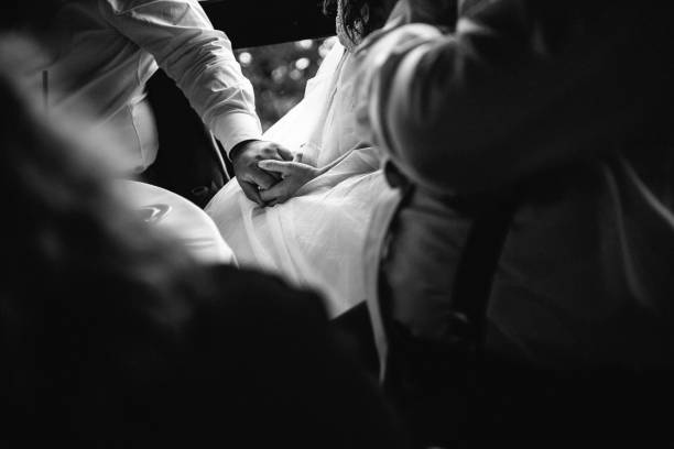 Artistic black and white photograph capturing an intimate moment at a wedding reception. Close-up shot of newlyweds holding hands, with the bride wearing a white wedding dress. The image conveys emotional connection and tenderness through minimalist composition, focusing on the couple's intertwined hands against the white fabric of the wedding dress. Soft lighting and shallow depth of field create a romantic atmosphere.