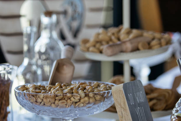 Elegant presentation of roasted cashew nuts served in a vintage cut crystal bowl at an upscale catering event or party. The composition includes blurred glass bottles in the background and additional serving platters, suggesting a sophisticated buffet setup. A decorative label holder adds a professional touch to the display.