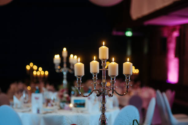 Silver candelabra with white pillar candles creating a romantic atmosphere at an elegant wedding reception. The five-arm candelabra stands prominently on a white-clothed table, with soft bokeh lights in the background. Purple and pink ambient lighting adds a modern touch to the classic decor. The setting suggests a formal indoor celebration with carefully curated table settings visible in the blurred background.