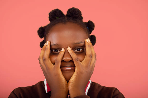 A playful portrait of a young woman with natural hair covering her face with her hands, leaving her eyes visible. Her expression adds a lighthearted, fun energy to the image, set against a coral backdrop.