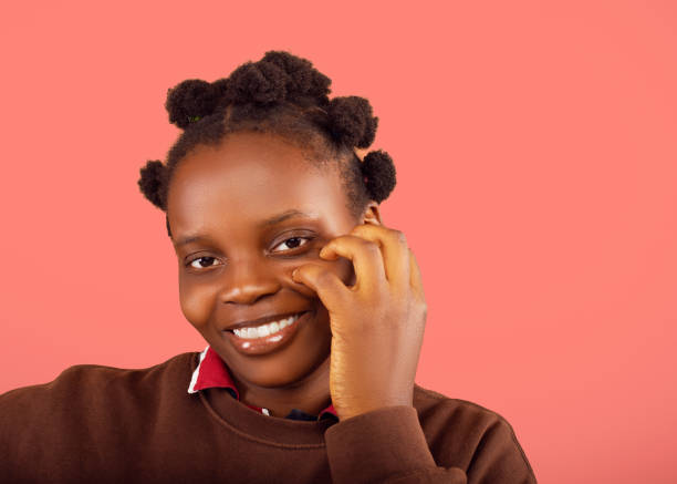 A joyful portrait of a young woman with natural hair, smiling brightly while gently holding her cheek. The image conveys a sense of happiness and warmth against a vibrant coral background.