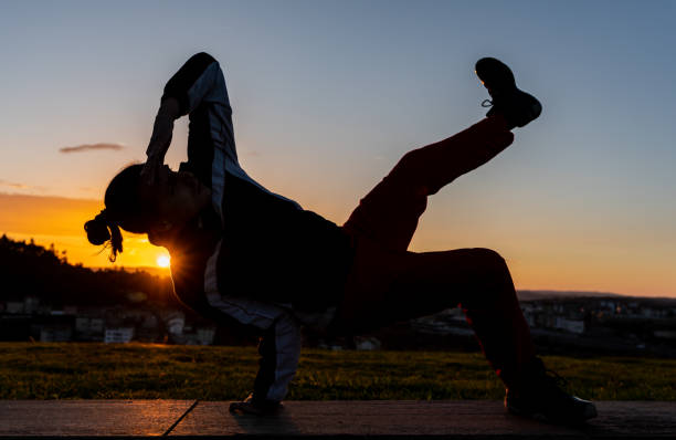 Backlit silhouette of a young man doing a breakdance position on a concrete ledge in front of a sunset with the city in the background. Serene and peaceful scene as the person is in a quiet and peaceful environment