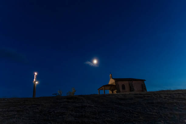istock Small hilltop chapel illuminated under a full moon with a glowing statue in the deep blue night sky. 2188016846