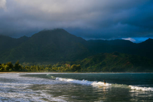 Vast shot of surfers riding waves in hanalei bay, with beautiful hawaiian mountains on the background. Taken in Kauai, Hawaii, during the day.