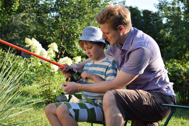 Man and boy standing with fishing rods on riverbank at sunset. Father focuses on fishing line while son holds rod, enjoying outdoor family activity near calm water.
