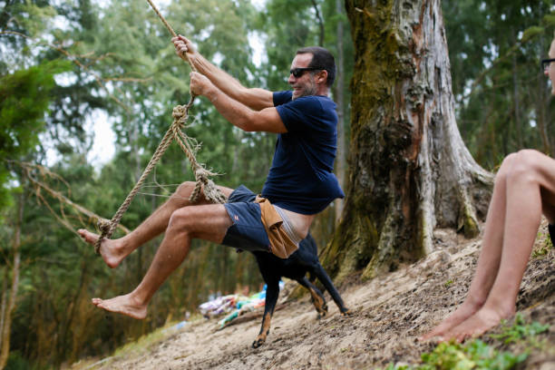 Shot of a middle aged man swinging on a rope in hawaiian forest, his son and dog near. Taken in Kauai, Hawaii, during the day.