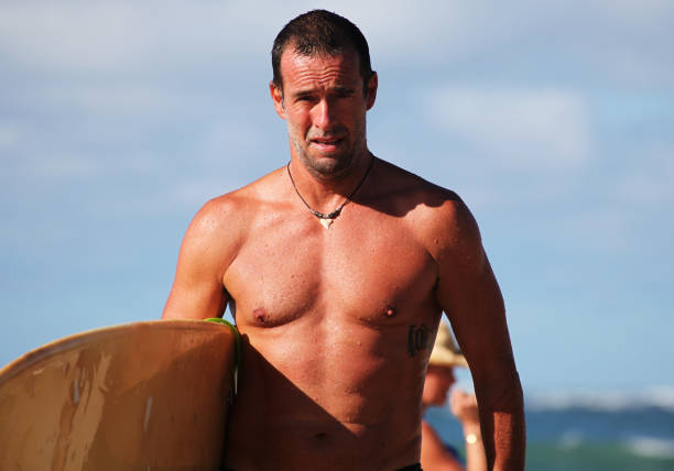 Shot of a fit man holding a surfboard coming out of the sea and walking toward the camera, sky and sea behind. Taken in Kauai, Hawaii, during the day.