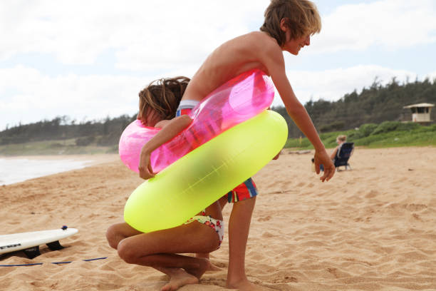 Shot of two siblings playing with flotation devices on hawaiian beach near the sea, sky and forest behind. Taken in Kauai, Hawaii, during the day.