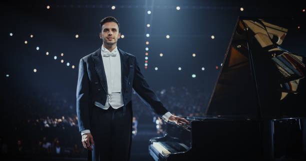 Classical Musician Posing for Camera in a Suit, Looking Straight and Smiling. Young Pianist Standing on a Concert Stage Next to Grand Piano with Excited Audience Sitting in the Background