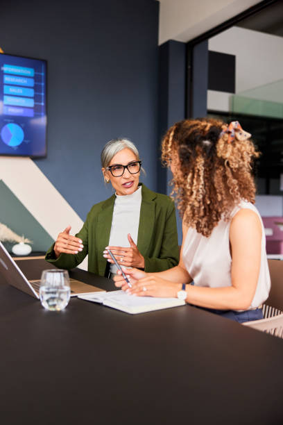 Mature female manager explaining and gesturing to young colleague at desk stock photo