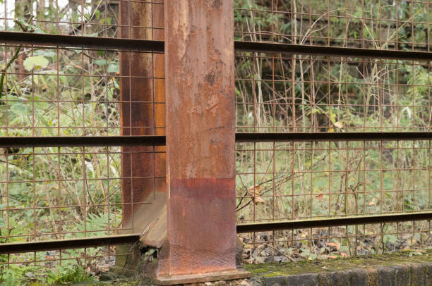 Close-up of a rusty metal beam supporting a wire mesh fence, with overgrown vegetation in the background, creating a scene of decay and abandonment
