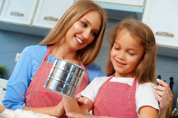 Middle-aged redhead mother serves dinner to her autistic teenage son and young daughter in their home kitchen. The 47-year-old woman wearing a colorful blue and yellow patterned blazer over a black top dishes food from a serving bowl onto a yellow plate. Her 18-year-old son in a black shirt sits at the table with a cookie on his plate while her 11-year-old daughter with long brown hair looks at her plate. The wooden kitchen features a table set with wine glasses, various dishes including what appears to be roast chicken, and a vase of yellow flowers. The scene captures a typical family dinner with focus on the mother's caregiving role.