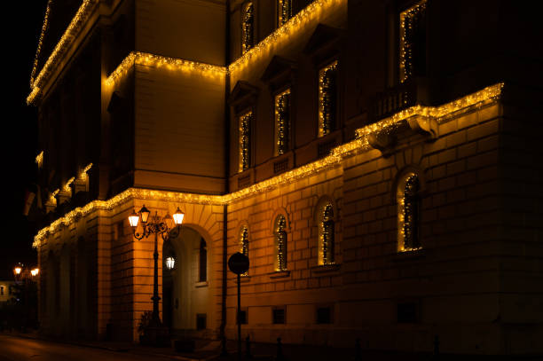 A corner view of an ornate building illuminated with warm, golden string lights for the festive season. The architectural details are highlighted by the glow, creating a cozy and celebratory nighttime scene.