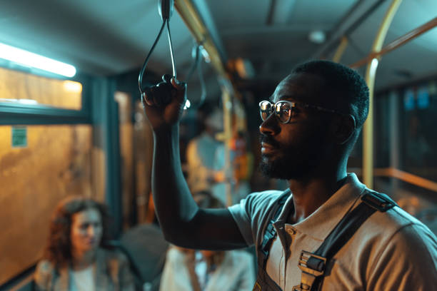 Black man in overalls holding onto a strap on a bus