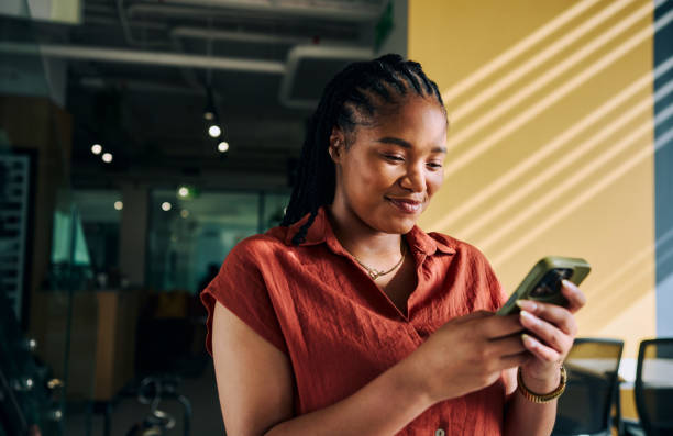 Young businesswoman using mobile phone, smiling and standing in modern office