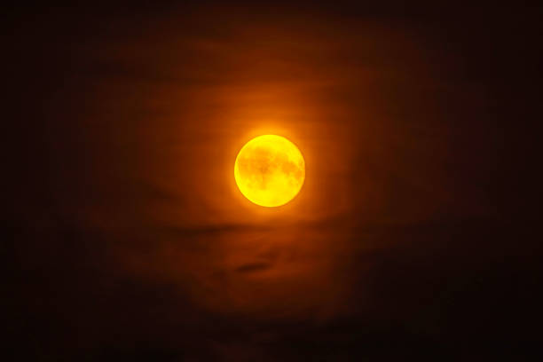 Bright full moon glowing in dark night sky surrounded by thin clouds with an orange hue.