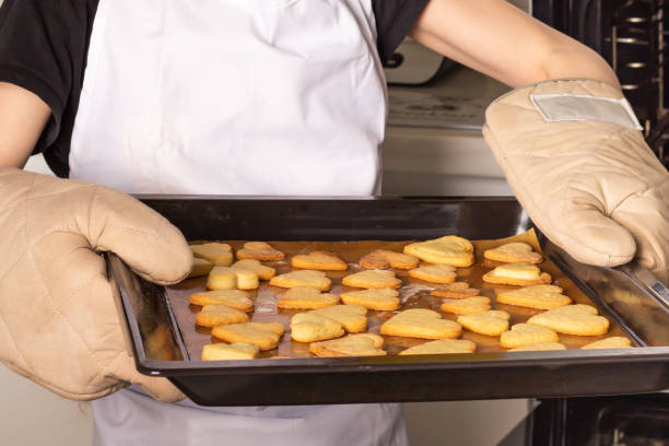 A person wears oven mitts and carries a baking tray filled with heart-shaped cookies freshly made in a warm kitchen. The atmosphere is inviting and homey, showcasing a love for baking.