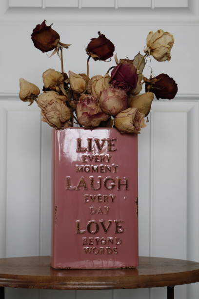Dried roses in a colorful square vase on a table. The vase has the phrase, Live every moment, Laugh every day, Love beyond words.