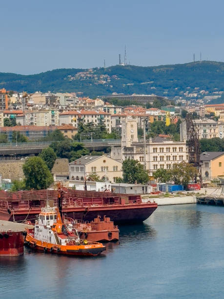 Trieste, Italy - June 29, 2024: Commercial harbor. Tugboat docked at drydock. Harbor management historic building with clock tower in back and cityscape behind. Hills on horizon