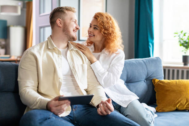 istock Cheerful loving spouses using digital tablet for shopping online, looking at each other and smiling, couple relaxing on couch in living room 2188100471