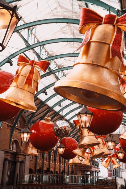 Christmas-themed decorations featuring oversized golden bells with red bows and bright red baubles hang from the ceiling at Covent Garden Market in London, UK. The festive scene showcases the iconic market's holiday transformation, with glittering ornaments and traditional lighting creating a warm and celebratory atmosphere. Captured during the 2024 holiday season, this image highlights the popular destination's charm and seasonal appeal.