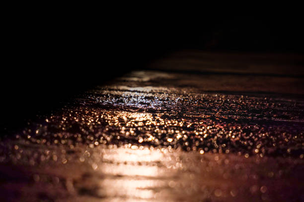 A close-up shot of wet pavement illuminated by soft, warm light in a dark environment. The bokeh effect highlights shimmering water droplets, creating a moody and artistic composition perfect for backgrounds, urban themes, or atmospheric designs. Ideal for use in creative projects or stock photography needs