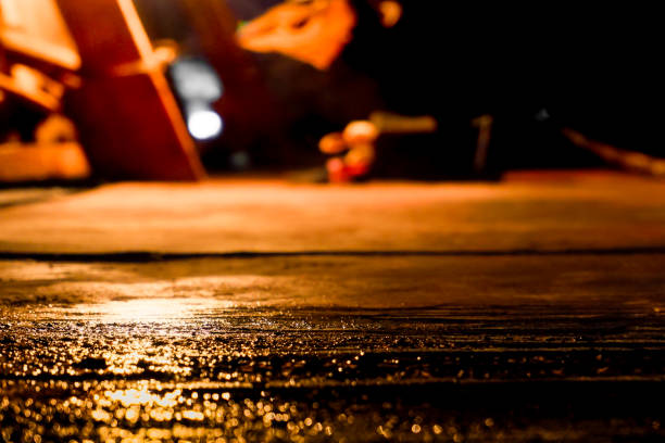 A close-up shot of wet pavement illuminated by soft, warm light in a dark environment. The bokeh effect highlights shimmering water droplets, creating a moody and artistic composition perfect for backgrounds, urban themes, or atmospheric designs. Ideal for use in creative projects or stock photography needs