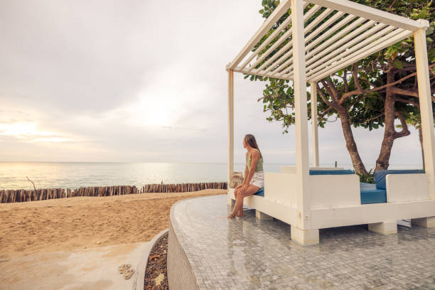 Woman Sitting on Beach Bench Looking at Ocean View Woman in a green top and white skirt sitting on a beach bench, gazing at the ocean. Serene beachfront setting with trees. pretty beach house stock pictures, royalty-free photos & images