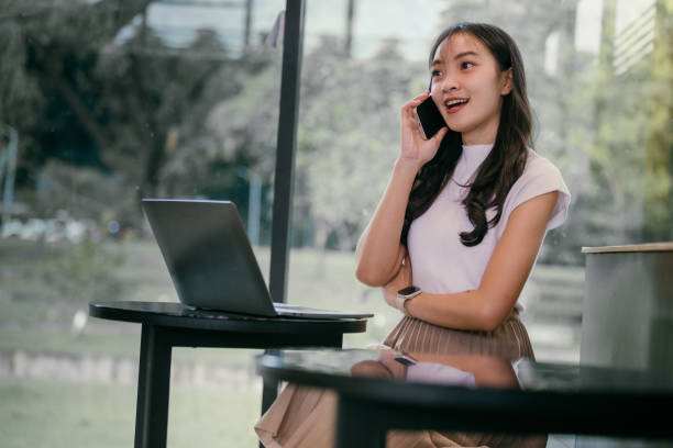 istock Young asian freelancer chatting on the phone while working on her laptop in a cozy cafe, relishing the warm natural light and inviting atmosphere around her 2188136349