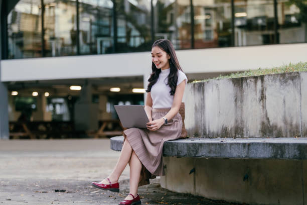 istock Young asian woman sitting on a bench outside a modern office building, working remotely on her laptop while smiling and enjoying the outdoor atmosphere 2188136533