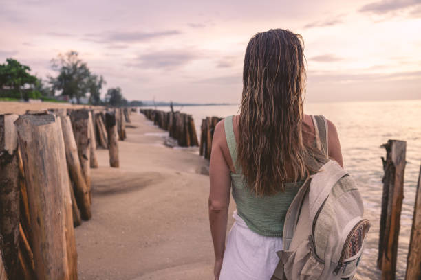 Woman in summer attire strolling on a sandy beach beside wooden posts. Serene sunset scene with an ocean view.
