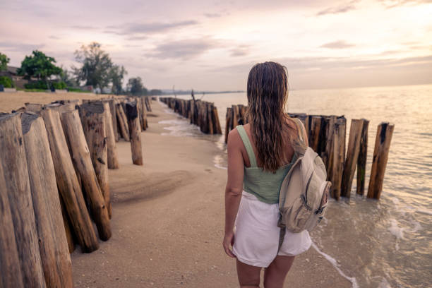 Woman in summer attire strolling on a sandy beach beside wooden posts. Serene sunset scene with an ocean view.