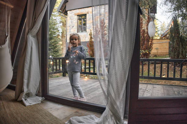 Cheerful blond haired kid in red checkered shirt looking at camera near opened window with view of snowy mountains and forest in hotel during vacation