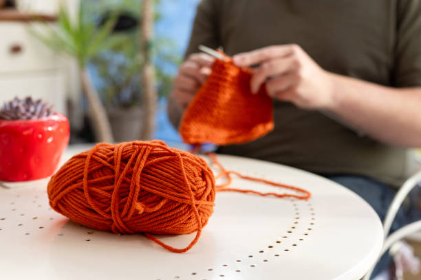 Close-up shot of an orange ball of wool on a table. An unrecognisable man is sitting and knitting using two knitting needles and orange wool.\n\nVideos are available similar to this scenario.