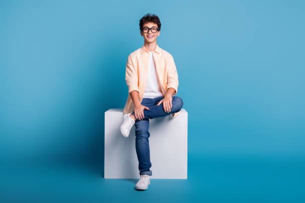 istock Smiling young man sitting stylishly on a white cube in front of a vibrant blue background, wearing casual and trendy fashion 2188188151