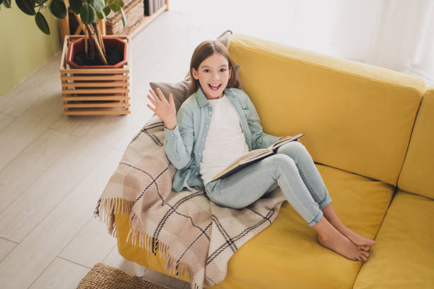istock Cheerful young girl relaxing on yellow sofa while reading a book indoors with daylight 2188189890