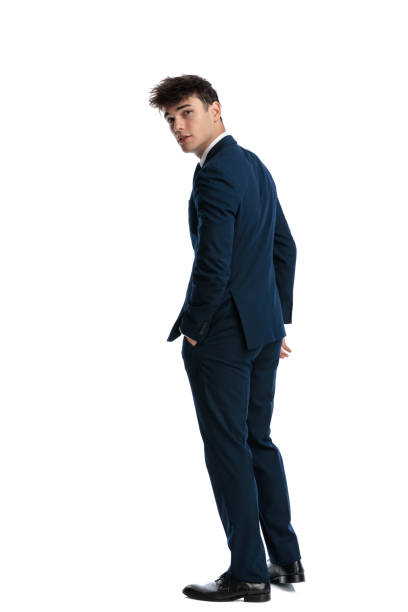 back view of cool young man in elegant navy blue suit with hand in pocket looking behind in front of white background