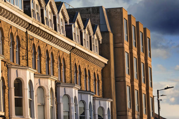 A row of brick townhouses with arched windows and bay windows, juxtaposed with a modern, light-brown multi-story building.  The scene is bathed in warm, late afternoon sunlight against a partly cloudy sky.
